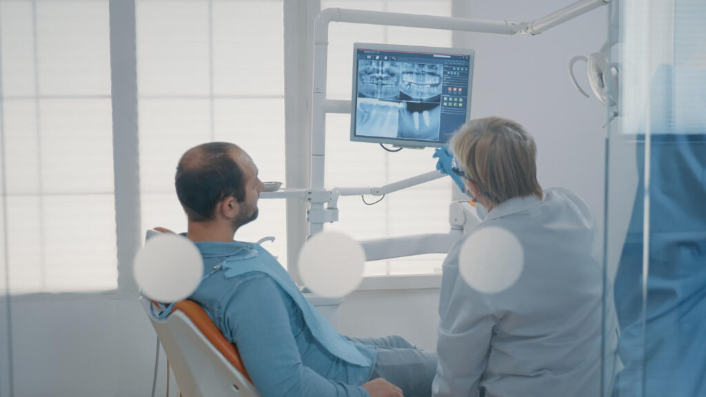 Un dentiste montre une radiographie dentaire panoramique sur un écran suspendu à son patient installé sur le fauteuil de soins.