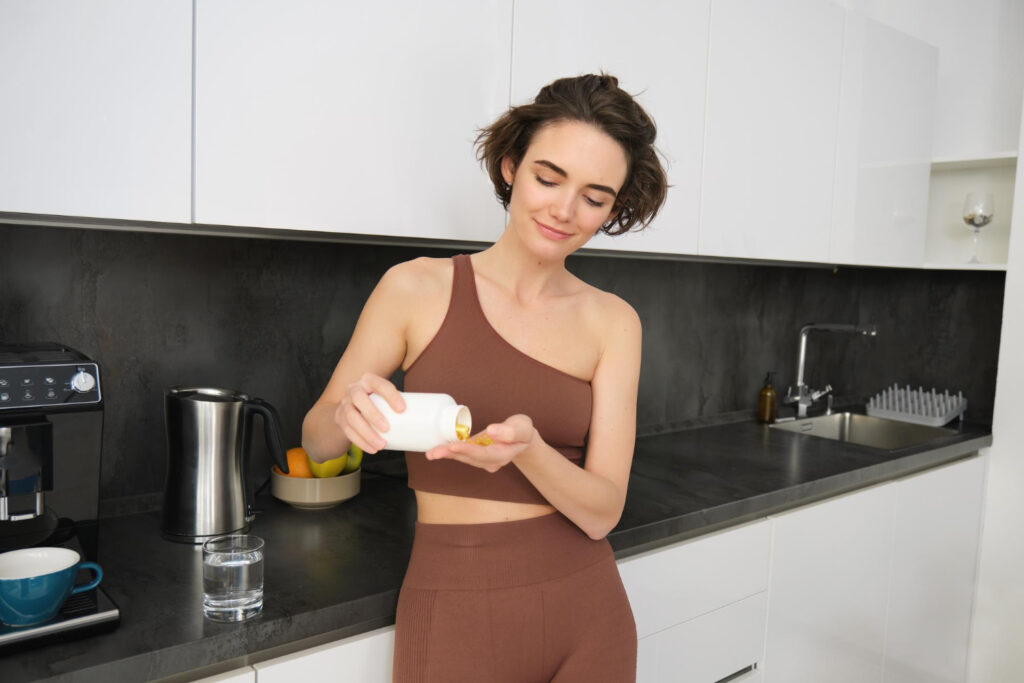 Une jeune femme en tenue de sport marron verse des compléments alimentaires dans sa main dans une cuisine moderne
