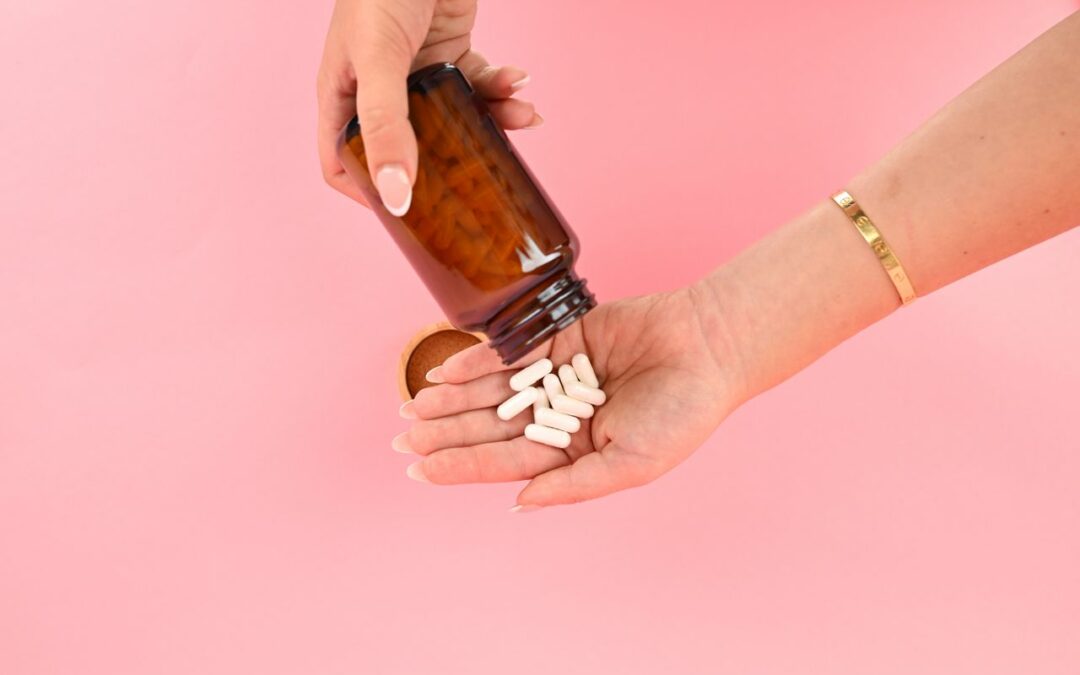 des mains versant plusieurs gélules blanches depuis un flacon en verre brun sur un fond rose uni