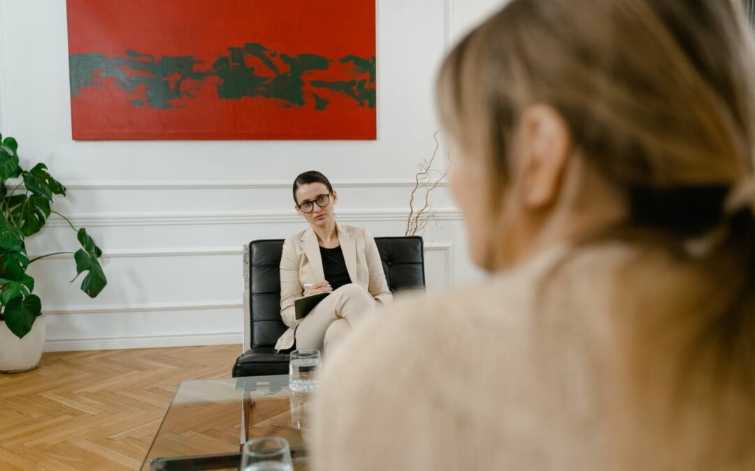 Une thérapeute en costume beige à l'écoute d'une cliente, vue en amorce depuis l'arrière de cette dernière, dans un bureau moderne décoré d'un tableau rouge.